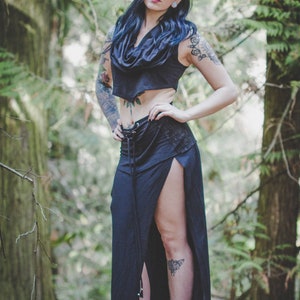 May include: A woman wearing a black crop top and a long black skirt with a side slit. She has dark hair and tattoos on her arms and legs. She is standing in a forest.