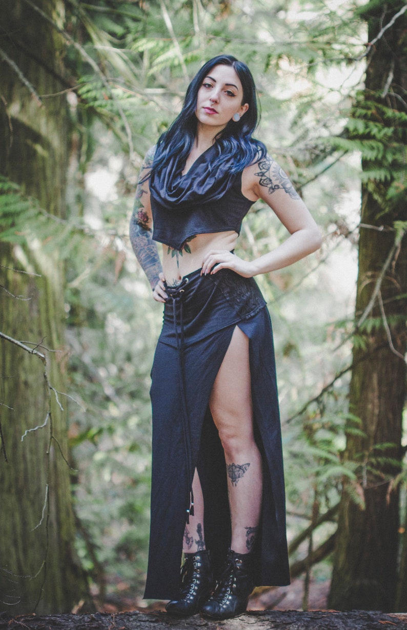 May include: A woman wearing a black crop top and a long black skirt with a side slit. She has dark hair and tattoos on her arms and legs. She is standing in a forest.