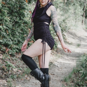 May include: A woman wearing a black, form-fitting, short-sleeved top with a gathered detail at the waist. She is wearing black knee-high socks and black combat boots. She has pink and purple hair and tattoos on her arms.