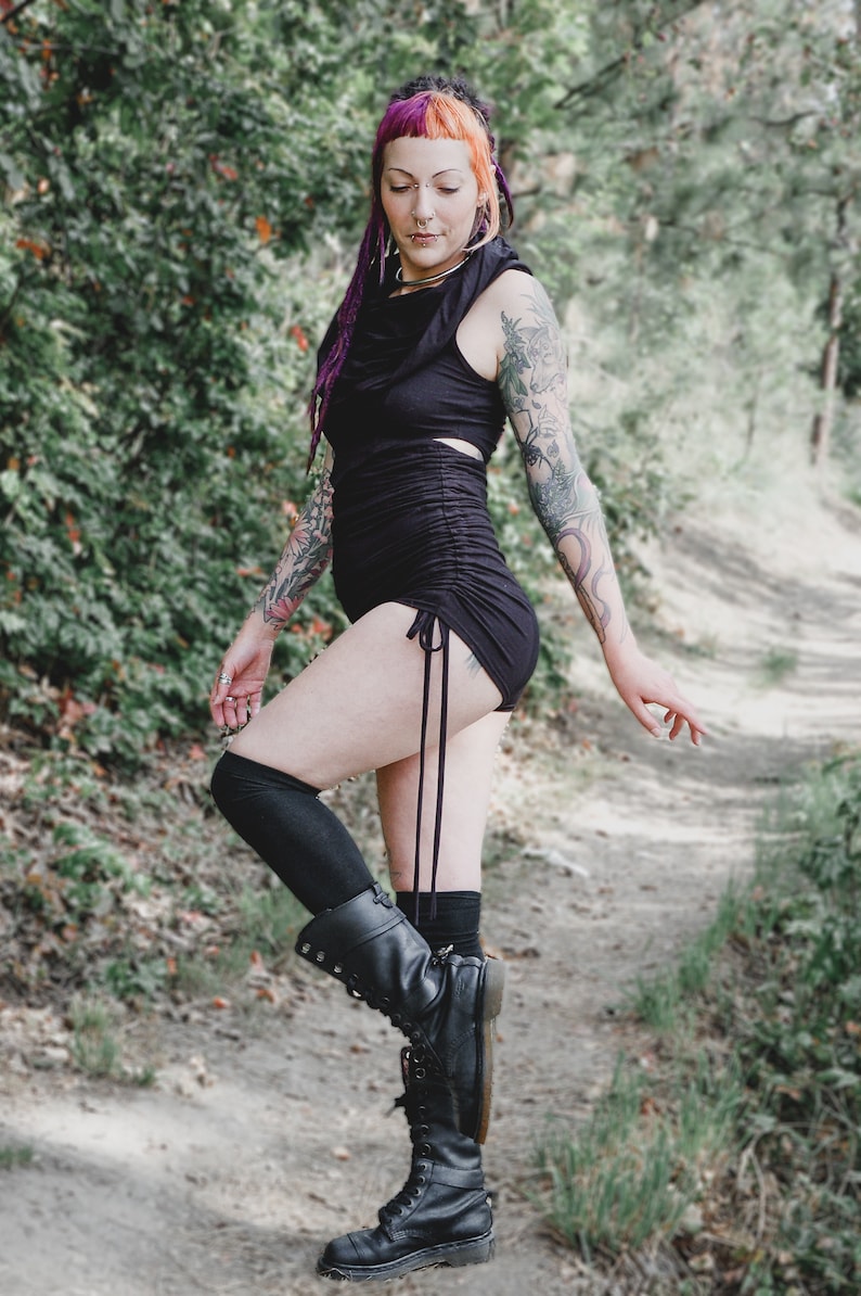May include: A woman wearing a black, form-fitting, short-sleeved top with a gathered detail at the waist. She is wearing black knee-high socks and black combat boots. She has pink and purple hair and tattoos on her arms.
