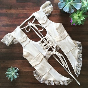 Peut inclure: Un gilet blanc en coton et dentelle avec une bordure en dentelle et un dos à lacets. Le gilet a des poches et des accents argentés.