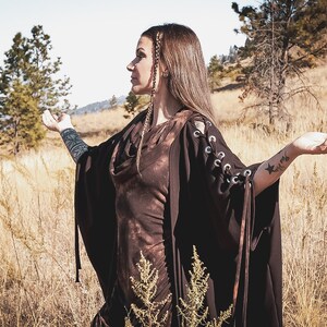 May include: A woman wearing a brown hooded dress with lace-up sleeves stands in a field of tall grass. She has long brown hair and is wearing a black choker necklace. The woman is looking to the right and has her arms outstretched.