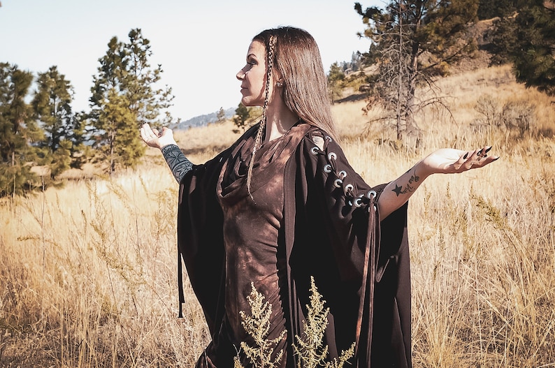 May include: A woman wearing a brown hooded dress with lace-up sleeves stands in a field of tall grass. She has long brown hair and is wearing a black choker necklace. The woman is looking to the right and has her arms outstretched.