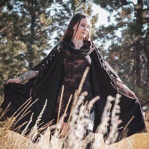 May include: A woman wearing a black hooded cloak with a tie detail, standing in a field of tall grass. The cloak has a tie detail at the neck and the sleeves are long and flowing.
