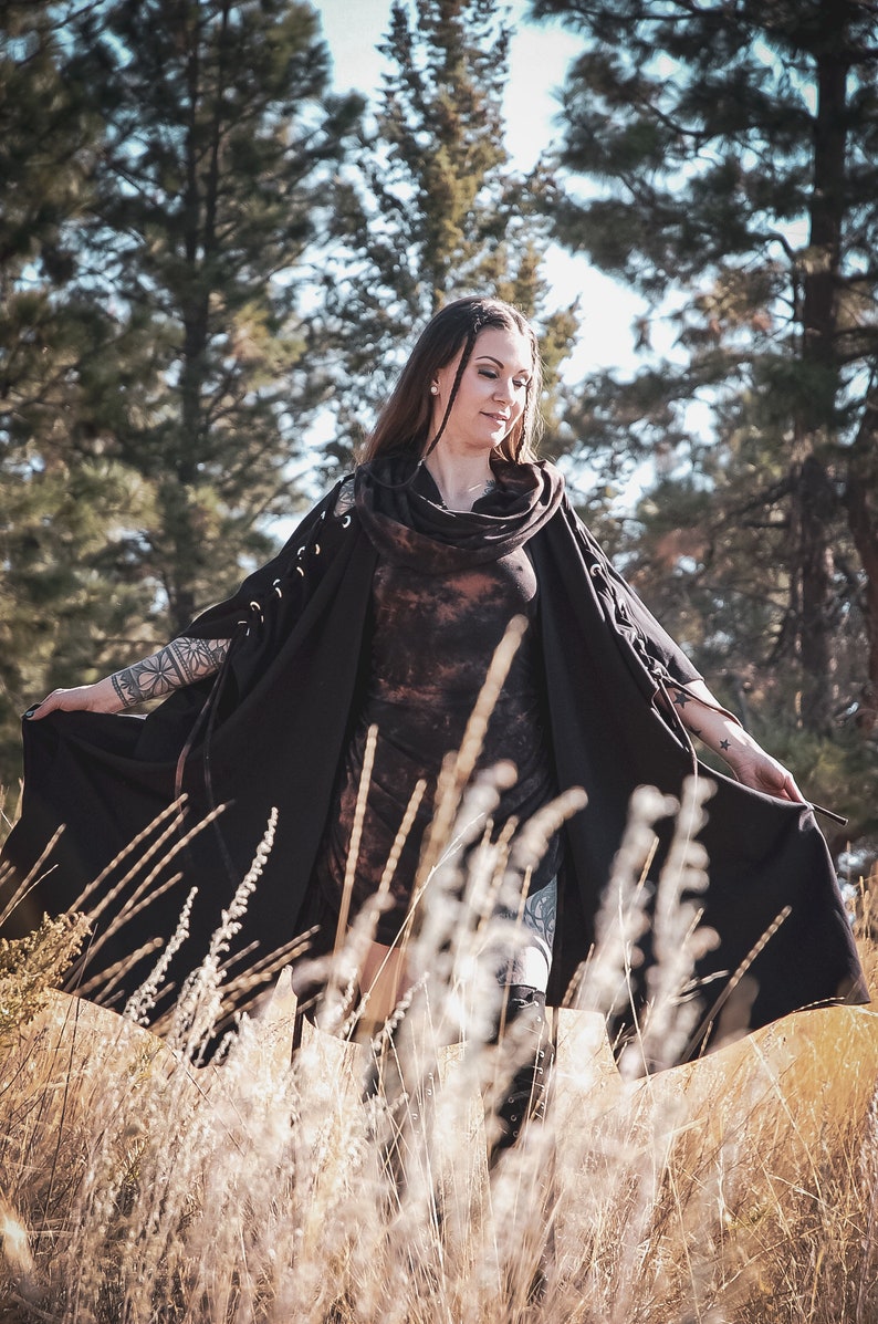 May include: A woman wearing a black hooded cloak with a tie detail, standing in a field of tall grass. The cloak has a tie detail at the neck and the sleeves are long and flowing.