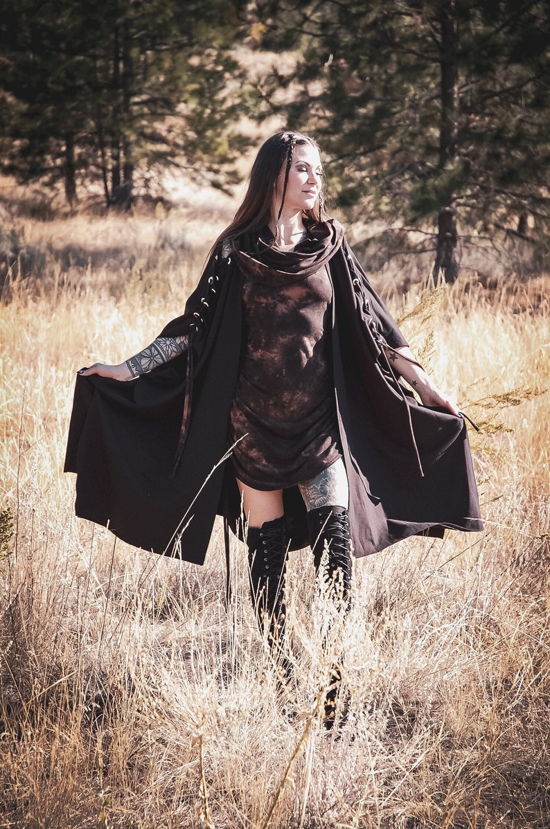 May include: A woman wearing a long, dark brown hooded cape with lace-up details. She is standing in a field of tall, dry grass. The cape has a tie at the neck and is flowing in the wind.