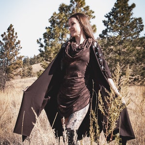 May include: A woman wearing a brown hooded dress with a long brown cape. She is standing in a field of tall grass with trees in the background.