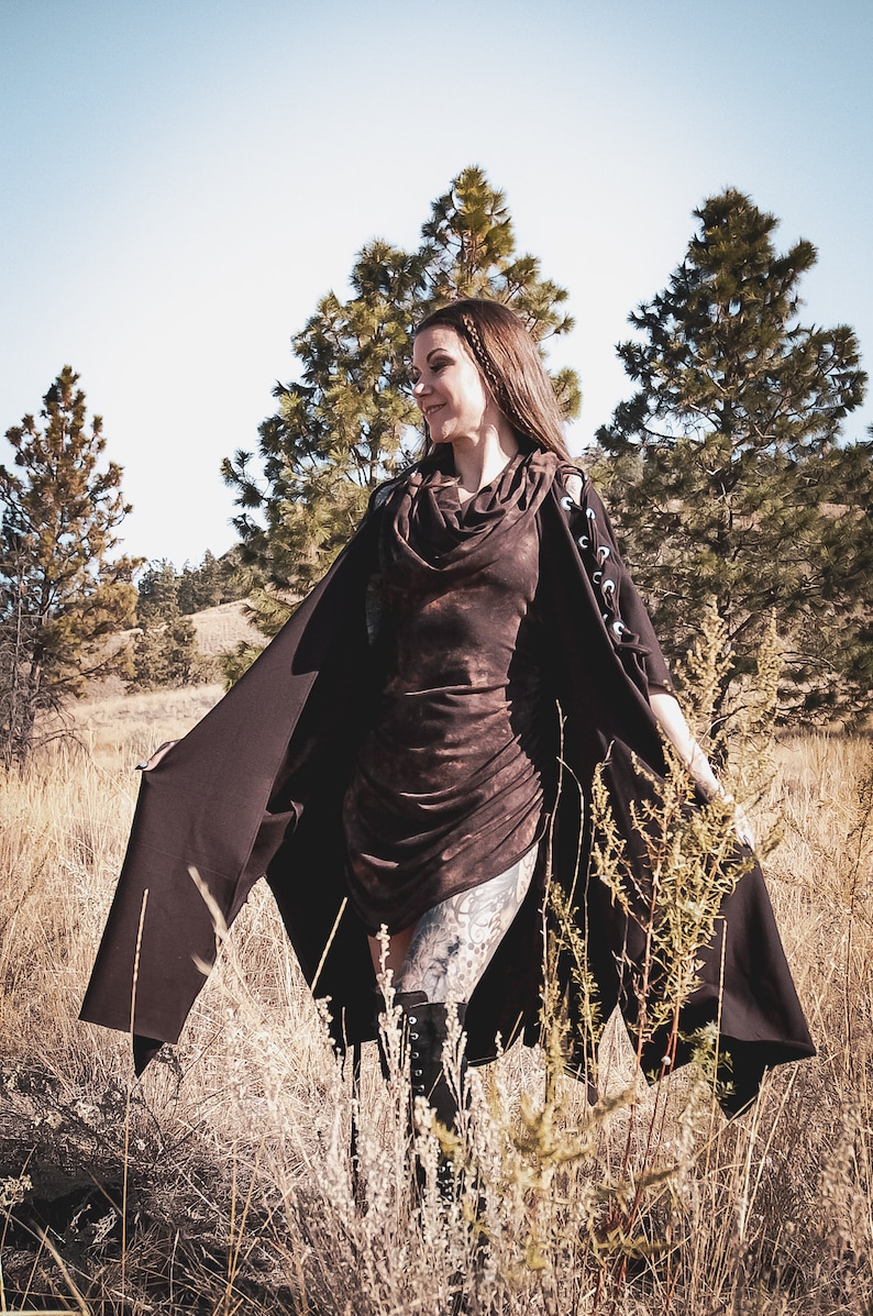May include: A woman wearing a brown hooded dress with a long brown cape. She is standing in a field of tall grass with trees in the background.