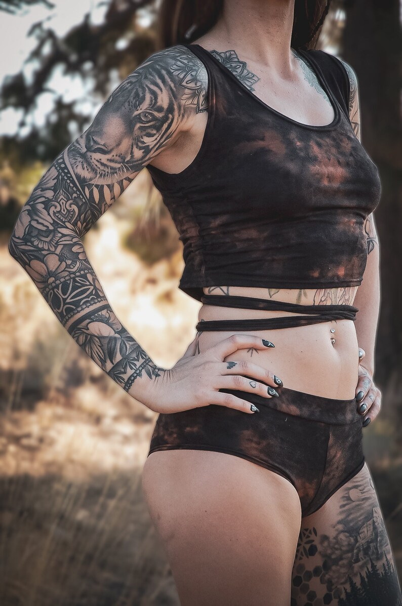 May include: A brown tie-dye two-piece outfit. The top is a cropped tank with a scoop neck. The bottoms are high-waisted shorts. The outfit is made of a soft, stretchy material. The model has tattoos on their arms and legs.