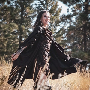 May include: A woman wearing a black hooded cape with lace detailing on the sleeves, standing in a field of tall grass. The cape is flowing in the wind, and the woman is smiling.