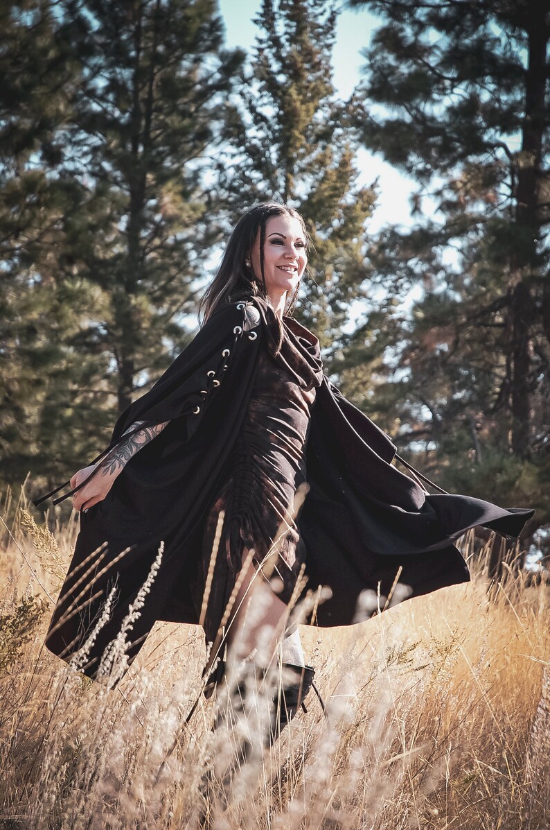 May include: A woman wearing a black hooded cape with lace detailing on the sleeves, standing in a field of tall grass. The cape is flowing in the wind, and the woman is smiling.