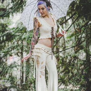 May include: A woman with purple hair and tattoos wears a white lace top and matching trousers with a floral design. She holds a white lace parasol with a floral pattern.