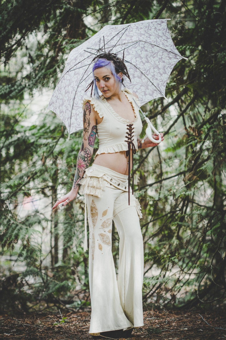 May include: A woman with purple hair and tattoos wears a white lace top and matching trousers with a floral design. She holds a white lace parasol with a floral pattern.