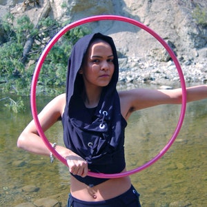 May include: A person wearing a black hooded top and a black skirt is holding a pink hula hoop in front of a river and rocky terrain.
