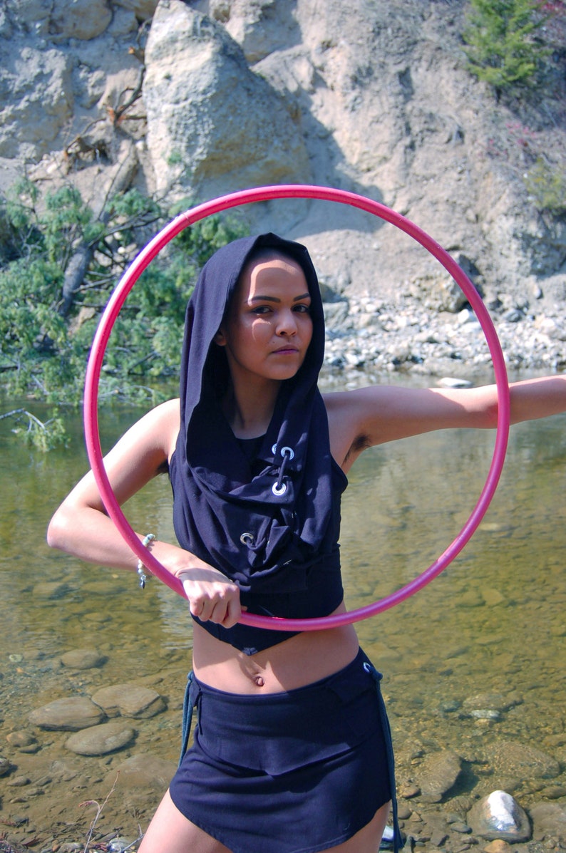 May include: A person wearing a black hooded top and a black skirt is holding a pink hula hoop in front of a river and rocky terrain.