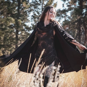 May include: A woman wearing a black hooded cape with a tie closure and a tie detail on the sleeves. She is standing in a field of tall grass.