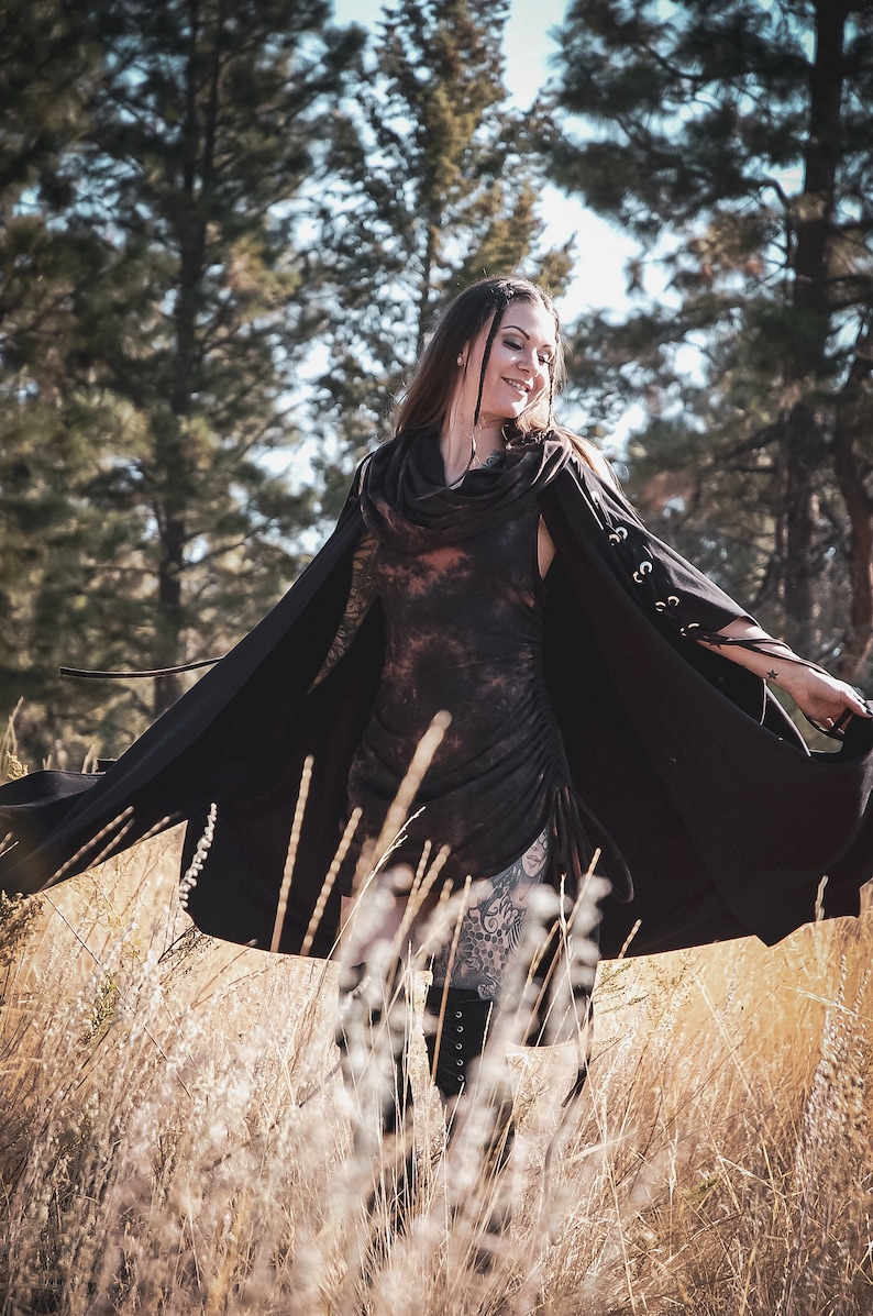 May include: A woman wearing a black hooded cape with a tie closure and a tie detail on the sleeves. She is standing in a field of tall grass.