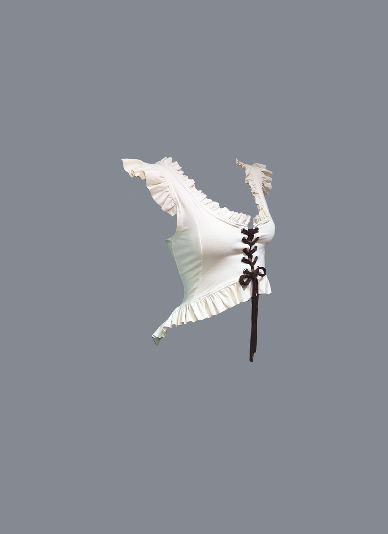 May include: A white crop top with a lace-up front and ruffled trim. The top has a brown ribbon tied at the front.