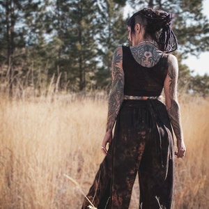 May include: A dark brown crop top and wide-leg trousers set. The trousers have a tie waist and a mottled pattern. The person in the image has tattoos on their arms and back. The background is a field of tall, dry grass.