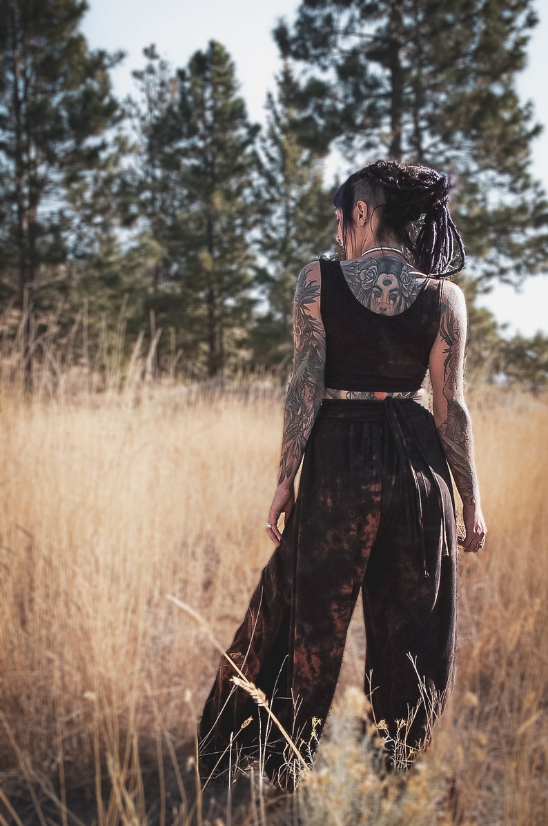 May include: A dark brown crop top and wide-leg trousers set. The trousers have a tie waist and a mottled pattern. The person in the image has tattoos on their arms and back. The background is a field of tall, dry grass.