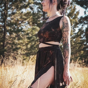 May include: A brown tie-dye crop top and skirt set. The sleeveless crop top has a wrap-around design, and the skirt features a high slit. The outfit is worn with black thigh-high socks. The background includes tall grass and trees.