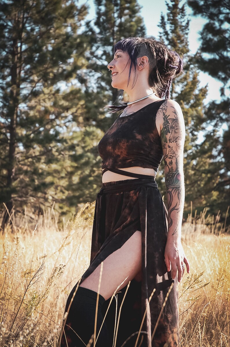 May include: A brown tie-dye crop top and skirt set. The sleeveless crop top has a wrap-around design, and the skirt features a high slit. The outfit is worn with black thigh-high socks. The background includes tall grass and trees.