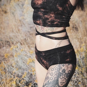 May include: Black tie-dye crop top and matching shorts set. The sleeveless top and high-waisted shorts feature a dark colour with reddish-brown accents. The outfit is tied at the waist with a matching fabric.
