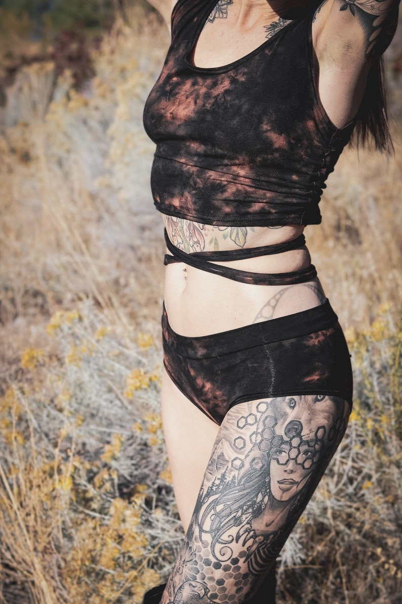 May include: Black tie-dye crop top and matching shorts set. The sleeveless top and high-waisted shorts feature a dark colour with reddish-brown accents. The outfit is tied at the waist with a matching fabric.
