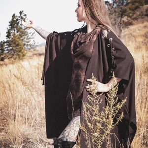 May include: A woman wearing a brown hooded cape with a tie closure and lace-up details on the sleeves. The cape is worn over a brown tie-dye top. The woman is standing in a field of tall grass.