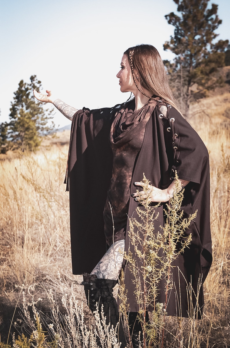 May include: A woman wearing a brown hooded cape with a tie closure and lace-up details on the sleeves. The cape is worn over a brown tie-dye top. The woman is standing in a field of tall grass.