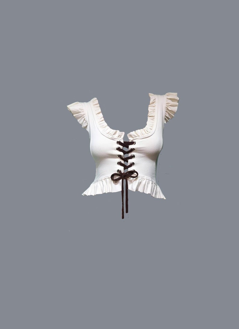 May include: A white crop top with a lace-up front and a ruffled neckline. The top has brown laces that tie into a bow at the centre.
