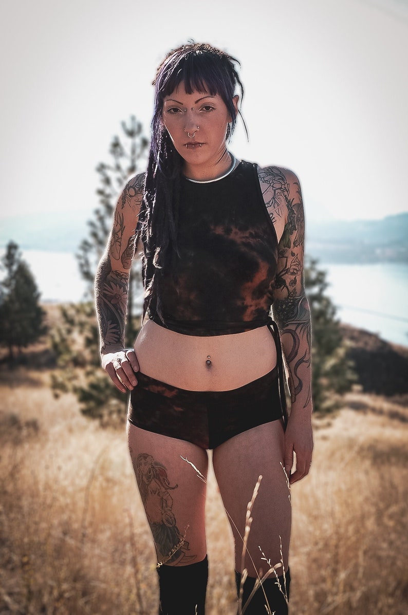 May include: A person with purple hair and tattoos is wearing a black tie-dye crop top and matching black tie-dye shorts. They are standing in a field with a body of water in the background.