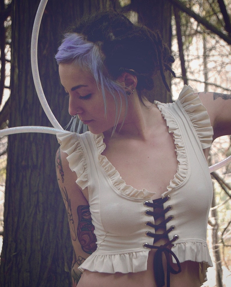 May include: A woman with purple and black hair is wearing a white, ruffled, lace-up crop top. She is standing in a woodland setting with trees in the background.
