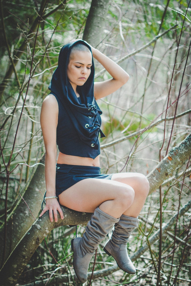 May include: A woman wearing a black hooded top with a lace-up front and a short black skirt. She is sitting on a tree branch with her legs crossed and wearing gray knee-high boots.