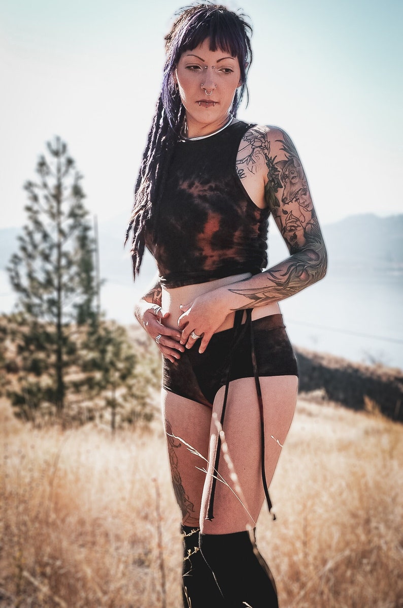 May include: A woman with purple hair and tattoos is wearing a black tie-dye crop top and matching shorts. The outfit has a tie detail at the waist. She is standing in a field of tall grass.