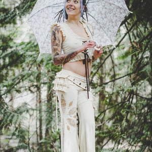 May include: A woman with purple hair and tattoos is wearing a white crop top and white trousers with lace detailing. She is holding a white umbrella with a floral pattern.