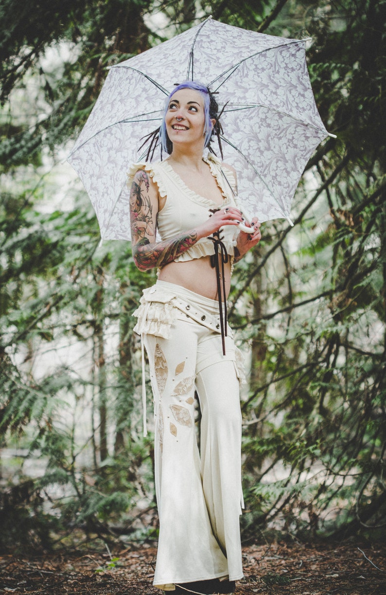 May include: A woman with purple hair and tattoos is wearing a white crop top and white trousers with lace detailing. She is holding a white umbrella with a floral pattern.