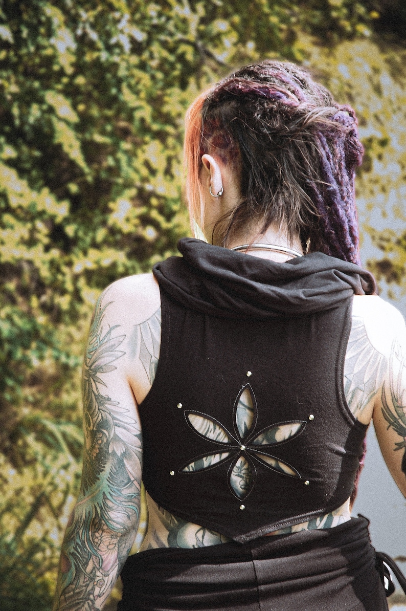 May include: A black sleeveless top with a cut-out design in the shape of a flower. The top has a hood and is worn by a person with tattoos on their arms.