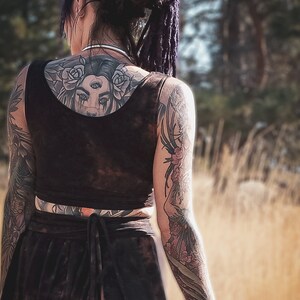 May include: A person with dark hair and tattoos, wearing a black crop top and skirt. The back tattoo features a woman's face with roses. The arms are covered in floral and other intricate tattoos. The outfit has a tie-waist detail.