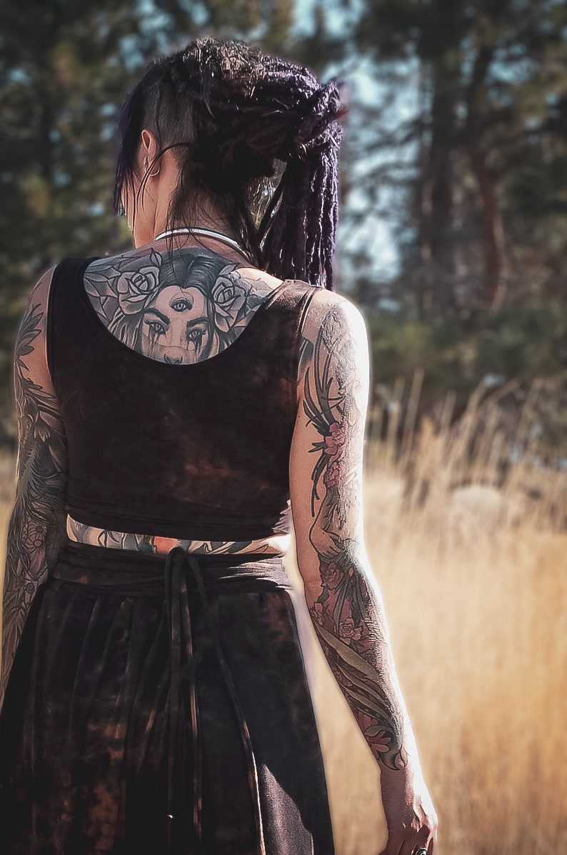 May include: A person with dark hair and tattoos, wearing a black crop top and skirt. The back tattoo features a woman's face with roses. The arms are covered in floral and other intricate tattoos. The outfit has a tie-waist detail.