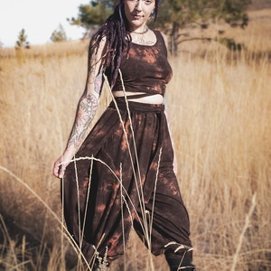 May include: A brown tie-dye crop top and harem pants set. The outfit has a unique tie-dye pattern with orange and brown hues. The model is standing in a field of tall, dry grass, wearing black boots.