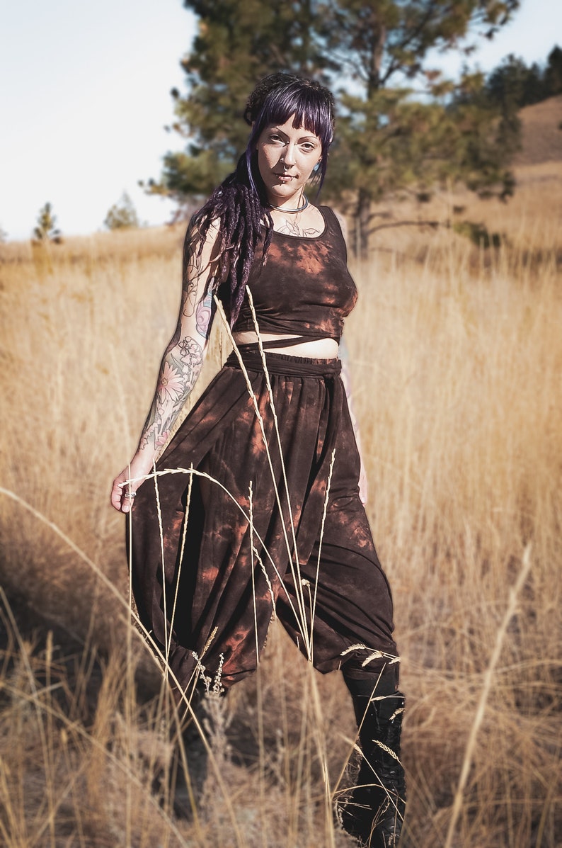May include: A brown tie-dye crop top and harem pants set. The outfit has a unique tie-dye pattern with orange and brown hues. The model is standing in a field of tall, dry grass, wearing black boots.