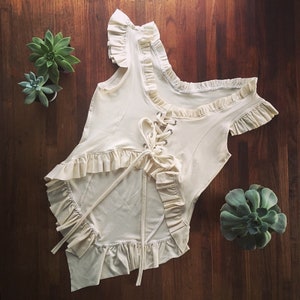 May include: A white cotton top with a lace-up front and ruffled trim. The top has a scoop neck and short sleeves.