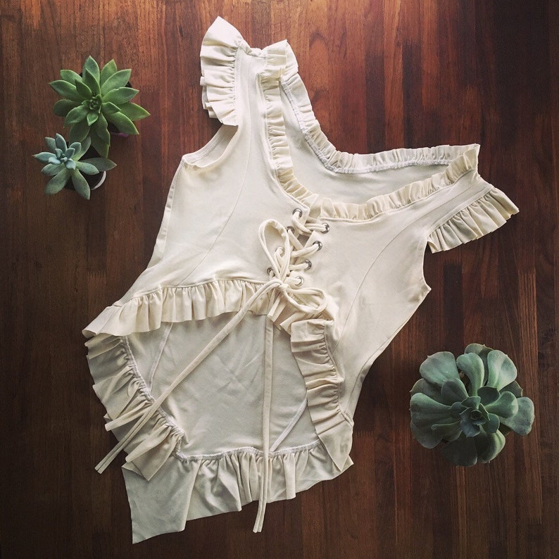 May include: A white cotton top with a lace-up front and ruffled trim. The top has a scoop neck and short sleeves.