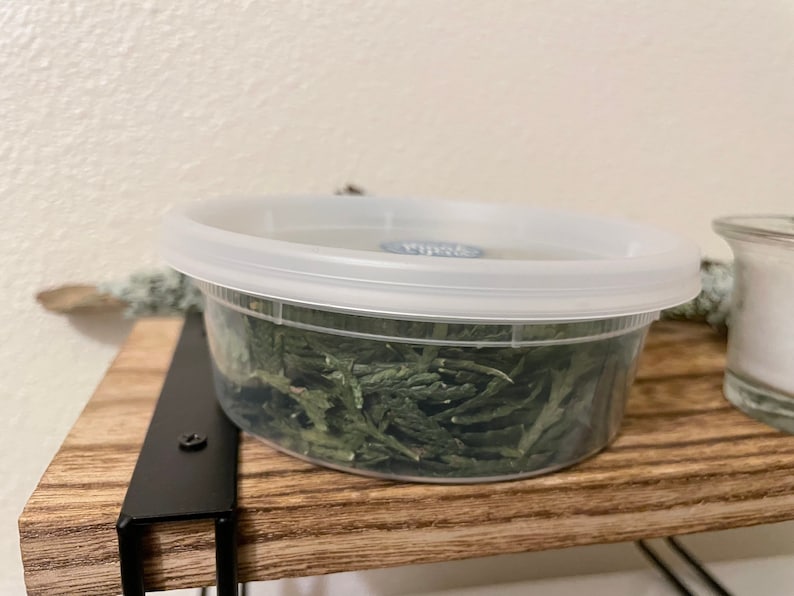 May include: A clear plastic container with a white lid, filled with dried green leaves. The container sits on a wooden shelf supported by a black metal bracket. A small glass jar is visible to the right.
