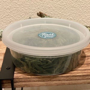May include: A clear plastic container with a white lid, filled with green dried leaves. A small blue and white sticker on the lid reads "Thank you". The container sits on a wooden shelf with black metal supports.