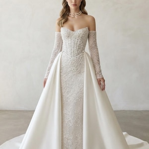 May include: A strapless white wedding gown featuring a corset-style bodice and long sleeves heavily embellished with intricate beadwork and pearls. The dress includes a smooth, floor-length overskirt with a train, paired with a pearl necklace and tiara.