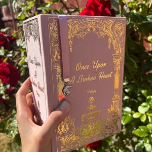 Pode incluir: Um livro rosa intitulado "Once Upon A Broken Heart" com detalhes decorativos dourados. O livro tem um marcador de fita rosa combinando com um pingente. O livro é segurado em frente a um fundo de rosas vermelhas e folhagem verde.