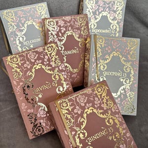 May include: A collection of hardcover books with floral and ornate gold foil designs. The books have titles such as "Releasing," "Taming," and "Binding 13." The covers are in shades of pink, gray, and gold, with matching ribbon bookmarks.