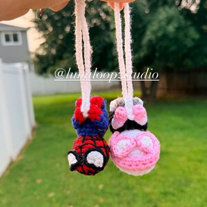 May include: Two small crocheted hanging ornaments with white yarn loops. One ornament features a red and blue superhero character with a spider web pattern and white eyes. The other is a pink, white, and black character with round ears.